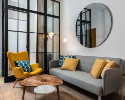 Colorful and cozy living room with a designer armchair and sofa along with a round decorative mirror and glass wall.